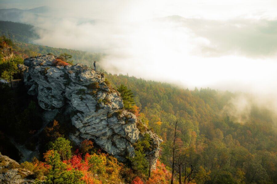 9 Hidden Gems to Visit in the South - 4 A person stands on the edge of a rocky cliff overlooking a forested valley with autumn foliage and low-lying clouds, discovering one of nature’s hidden gems.