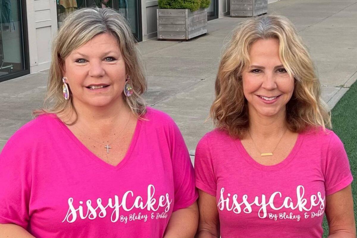 Two women with blonde hair wear matching bright pink "SissyCakes By Blakey & Dobie" T-shirts, standing outdoors on a Louisville sidewalk, ready to share their delicious sweets with the community.