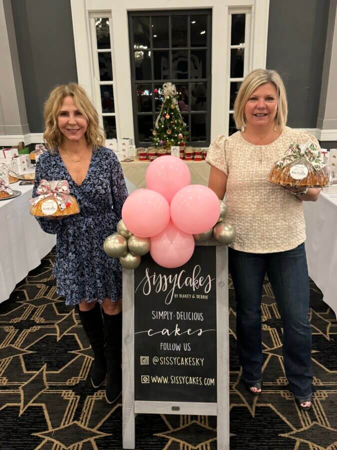 Meet the Sisters Behind Louisville’s SissyCakes Bakery - 2 Two women stand beside a SissyCakes sign in Louisville, holding baked goods with the famous Signature Glaze, while a festive Christmas tree and a tempting display of treats are visible in the background.