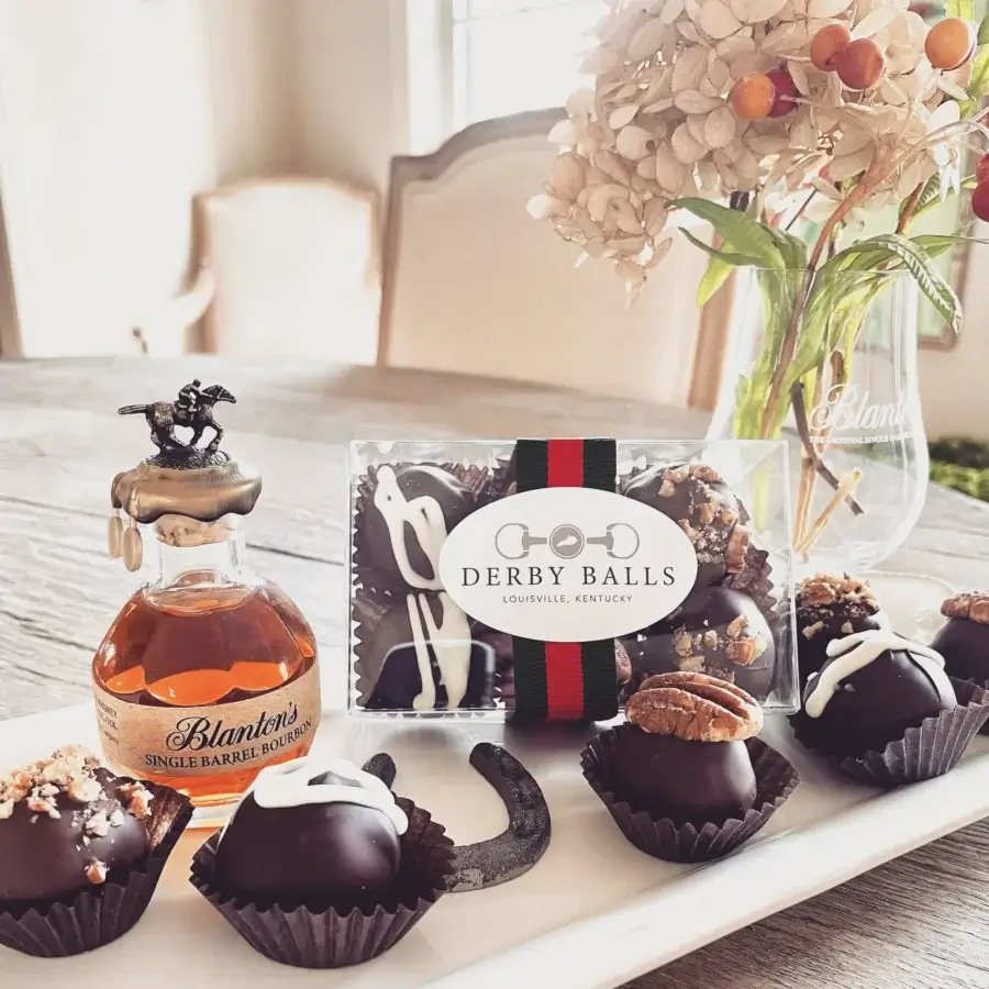 16 Louisville FINDS for April: Spring Style, Derby-Ready Decor & Must-Have Gifts - 11 A bottle of Blanton’s bourbon, a glass, Derby Balls chocolates, assorted truffles, a horseshoe, and spring flowers on a dining table capture the spirit of Louisville and its unique finds.