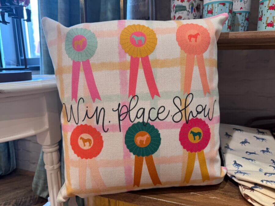 16 Louisville FINDS for April: Spring Style, Derby-Ready Decor & Must-Have Gifts - 8 A decorative pillow featuring colorful award ribbons and the text "Win: Place: Show," perfect for adding a touch of Spring charm to your Louisville Finds, displayed on a wooden shelf.