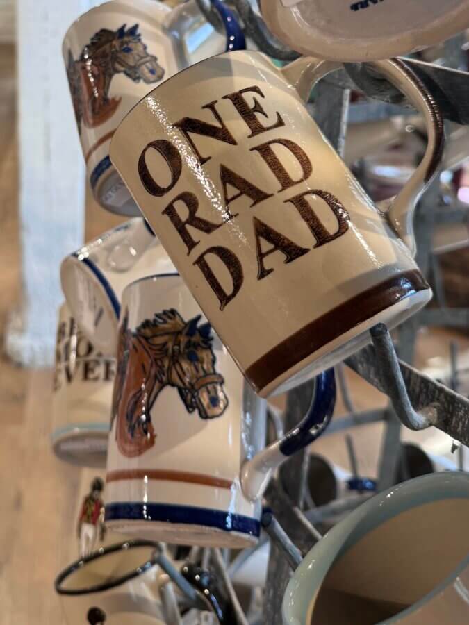 16 Louisville FINDS for April: Spring Style, Derby-Ready Decor & Must-Have Gifts - 14 A ceramic mug on a display rack features the text "ONE RAD DAD" in bold letters, perfect for spring finds in Louisville, and is surrounded by other mugs with charming horse illustrations.