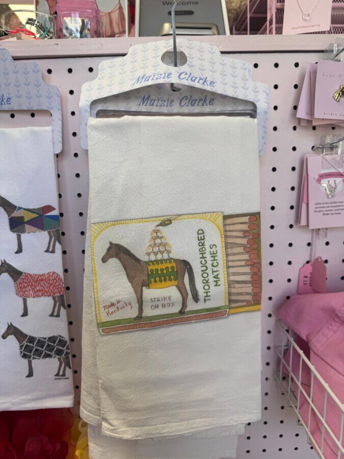 16 Louisville FINDS for April: Spring Style, Derby-Ready Decor & Must-Have Gifts - 13 A white tea towel features an illustration of a horse, a box of matches, and the text “THOROUGHBRED MATCHES” and “STRIKE ON BOX.” A perfect Louisville-inspired spring find, displayed on a pegboard rack.