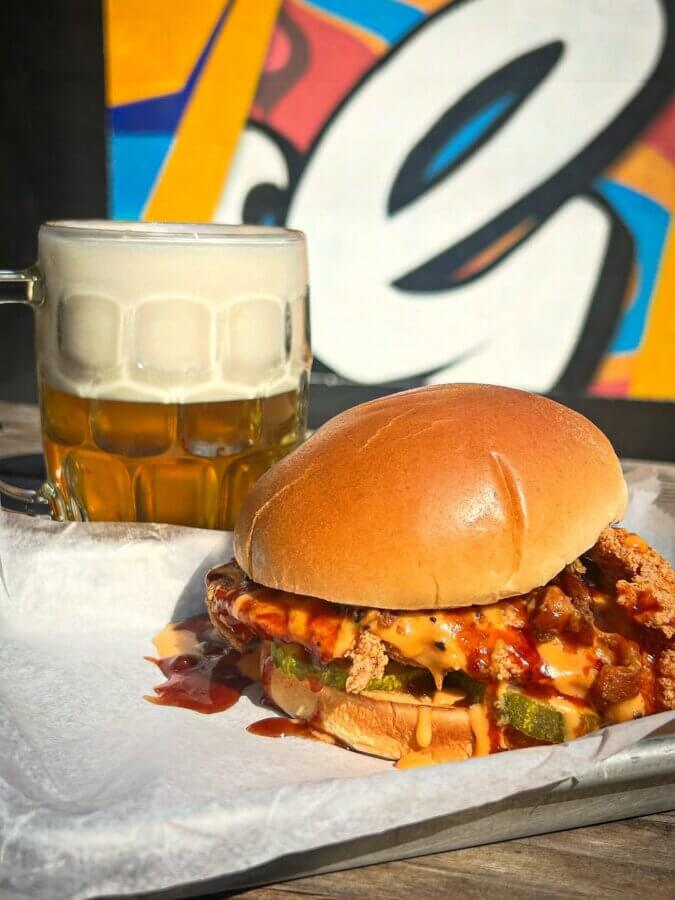 4 New Louisville Restaurants (+2 New Menus!) to Try This Month - 7 A fried chicken sandwich with sauce and pickles sits on a tray next to a mug of beer, with a colorful mural in the background.
