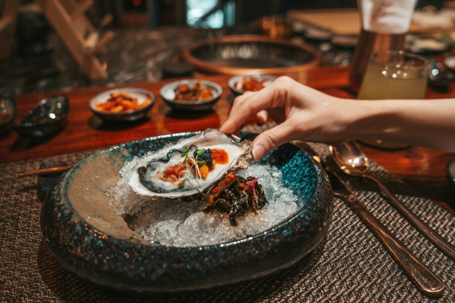 4 New Louisville Restaurants (+2 New Menus!) to Try This Month - 5 A hand holds an oyster topped with garnish over a bed of ice, with other small dishes and utensils on a table in the background.