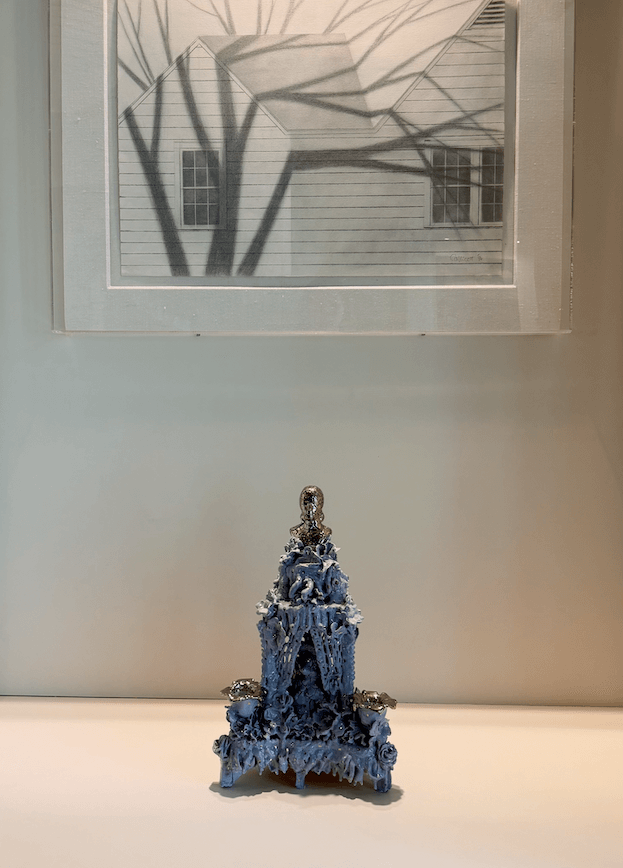 7 Memphis Galleries Exhibiting at Art by Design 2026 - 3 A blue sculptural object is displayed on a white surface beneath a framed black-and-white drawing of a house, featured in the upcoming Art by Design 2026 art exhibition at renowned Memphis galleries.