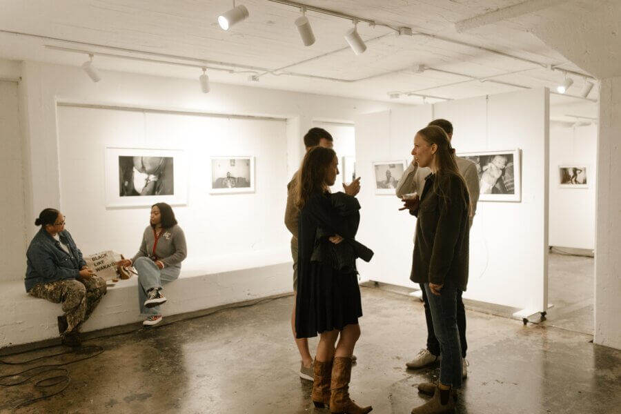 7 Memphis Galleries Exhibiting at Art by Design 2026 - 7 People stand and converse in a white-walled art gallery, viewing framed black-and-white photographs on display at the Art by Design 2026 exhibition. Two people sit chatting on a bench in the background of this Memphis galleries event.