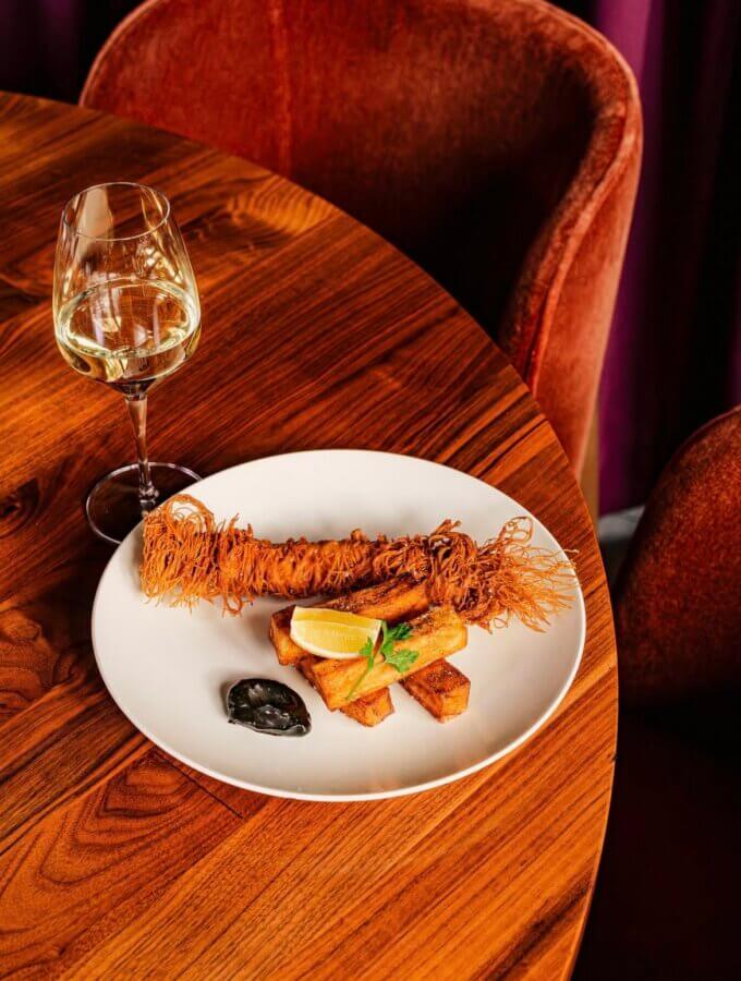 5 New Memphis Restaurants to Try This Month (+ What's Coming Soon!) - 12 A plated gourmet dish with a breaded, crispy item, rectangular potato pieces, a lemon wedge, black sauce, and a glass of white wine on a wooden table.