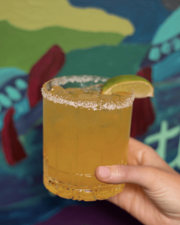 5 New Memphis Restaurants to Try This Month (+ What's Coming Soon!) - 11 A hand holds a glass of orange cocktail with a salted rim and a lime wedge garnish, against a colorful painted background.