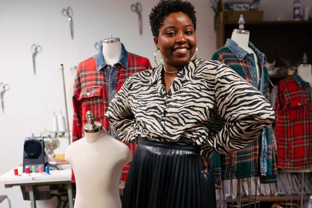 A woman stands smiling with hands on hips in Cari Harris’s sewing studio, surrounded by dress forms, sewing machines, and plaid shirts on mannequins.