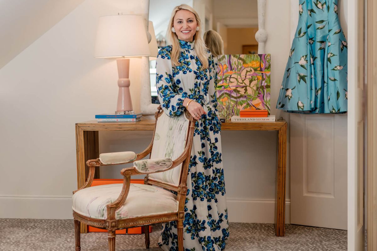 A woman in a long floral dress stands by a wooden desk with a lamp, books, and colorful artwork from the FACES collection in a well-decorated Meade Carlisle room.
