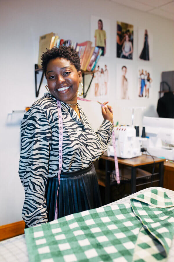 She's Changing the Way Memphis Thinks About Clothing - 3 A woman wearing a zebra print top and pleated skirt stands in a sewing room with a measuring tape around her neck, smiling at the camera.