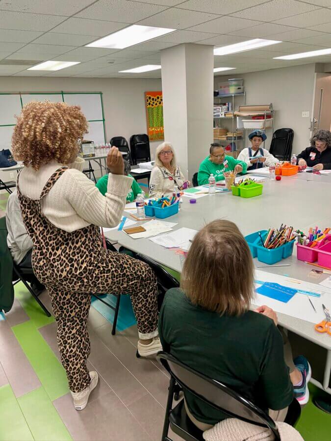 She's Changing the Way Memphis Thinks About Clothing - 5 A group of adults sit around tables covered with art supplies, while Cari Harris, in leopard-print overalls, stands and speaks to the group in a classroom setting.