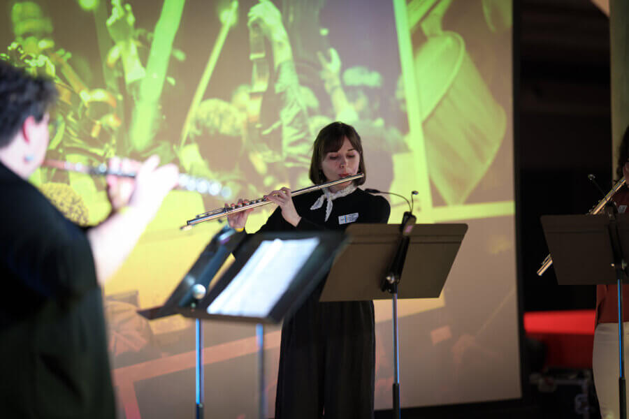 A woman plays the flute on stage with sheet music in front of her; two other musicians with flutes, including Jenny Davis, are partially visible, with a large projected image of FACES in the background.