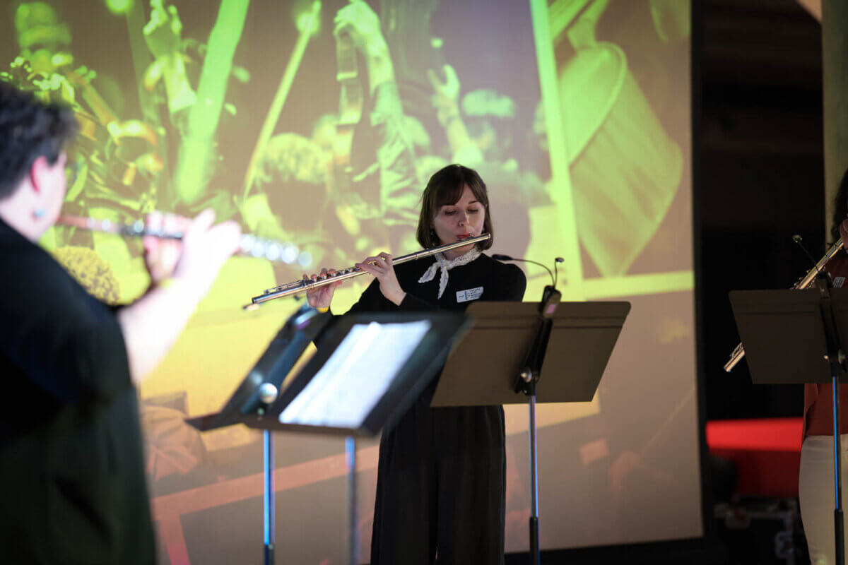 A woman plays the flute on stage with sheet music in front of her; two other musicians with flutes, including Jenny Davis, are partially visible, with a large projected image of FACES in the background.
