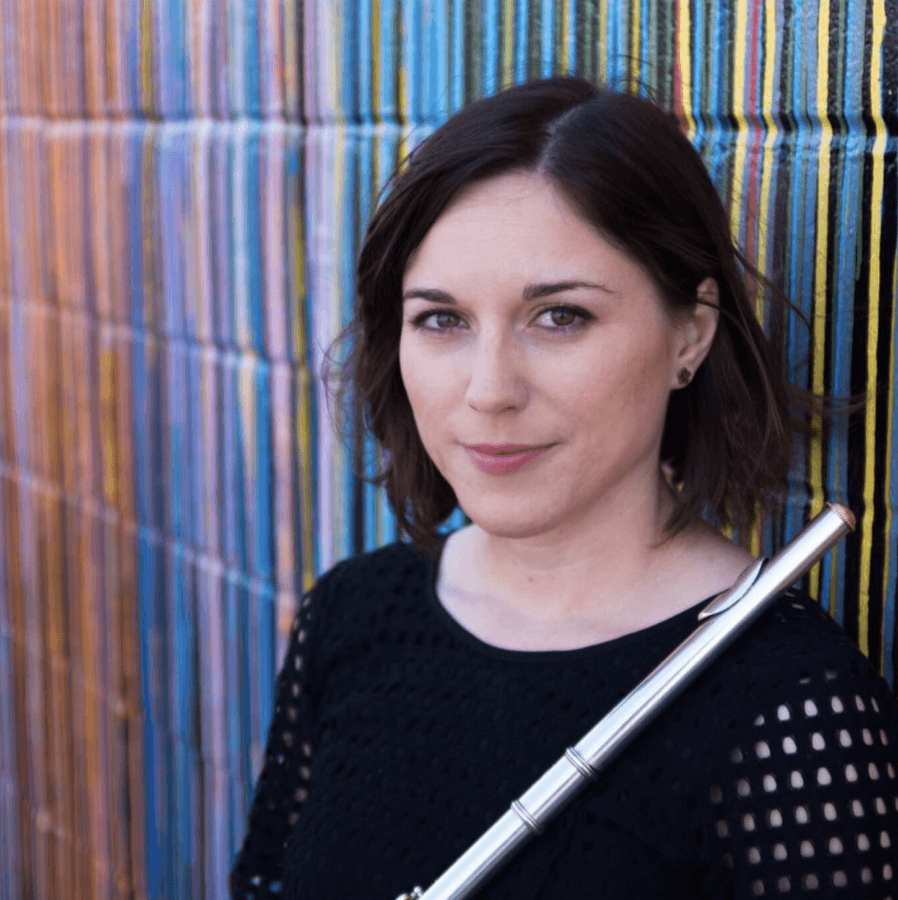 Meet the Local Flutist Behind the Memphis Youth Symphony Program - 2 Woman with short brown hair wearing a black top holds a flute against her shoulder, standing in front of a colorful, striped wall.