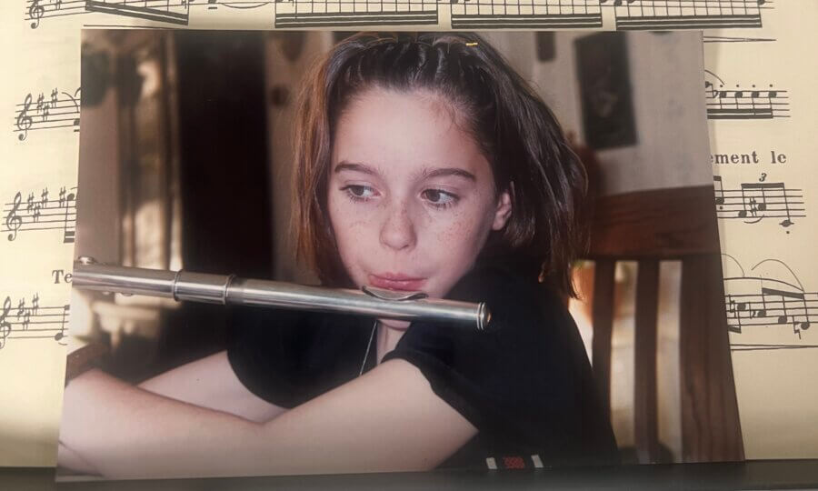 Meet the Local Flutist Behind the Memphis Youth Symphony Program - 3 A young person with freckled cheeks holds a flute near their mouth, seated indoors, with sheet music in the background.