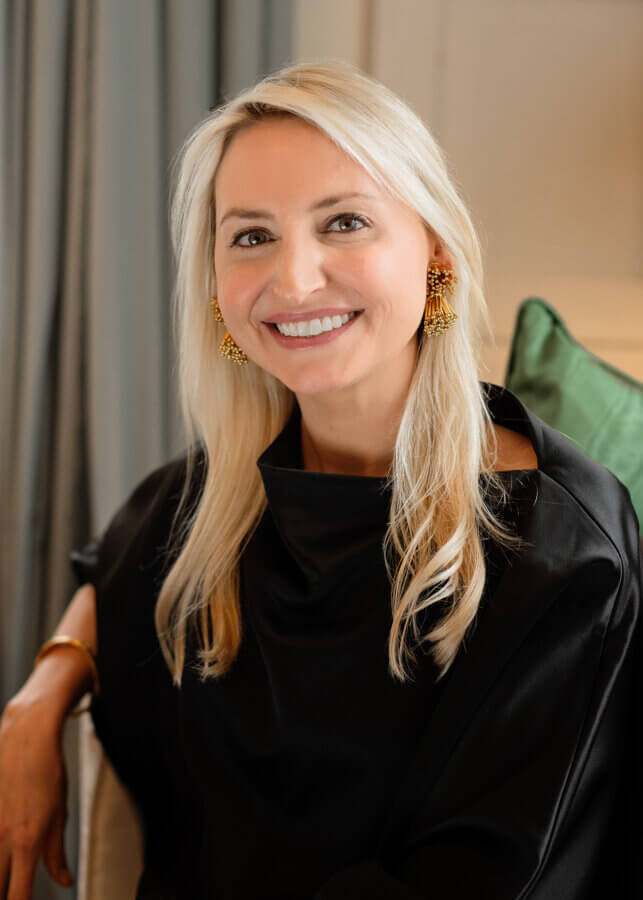 A woman with long blonde hair, wearing a black top and gold earrings, smiles while sitting on a chair with a green cushion in the background.