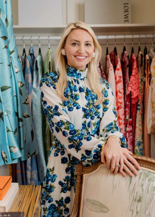 A woman in a floral dress stands by a chair in a Meade Carlisle boutique, with colorful dresses hanging on a rack behind her.