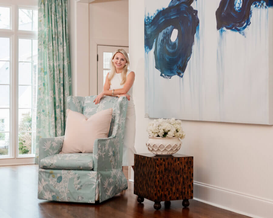 A woman stands smiling behind a patterned armchair in a bright living room with a large abstract painting, a floral arrangement, and floor-to-ceiling windows.