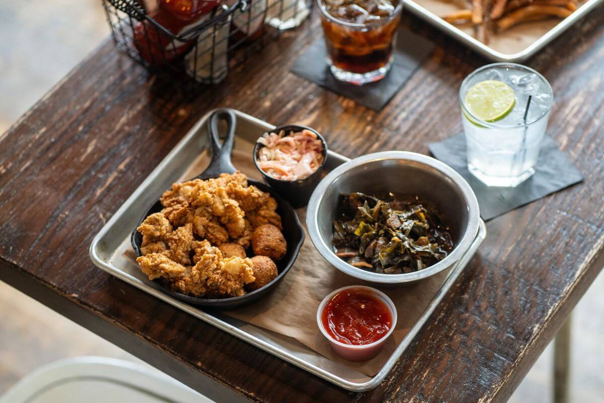 A tray at one of the top restaurants in Memphis featuring fried chicken, hush puppies, collard greens, coleslaw, ketchup, iced cola, and water with lime sits on a wooden table.
