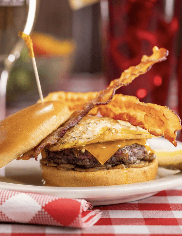 7 Must-Try Memphis Smashburgers - 4 A cheeseburger topped with crispy bacon and a fried egg on a toasted bun, served on a white plate with a pickle spear.