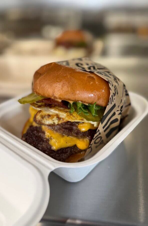 7 Must-Try Memphis Smashburgers - 2 A cheeseburger with lettuce, pickles, cheese, bacon, and a fried egg in a toasted bun, served in a takeout container.