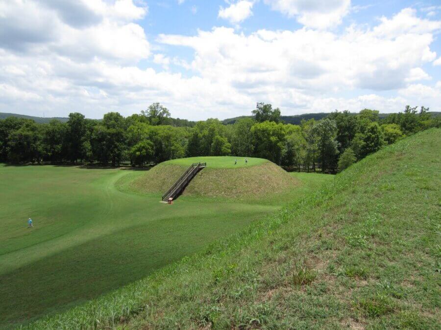 Hidden History in the Southβs Most Beautiful State Parks - 3 Grassy earthen mound with wooden stairs in an open field, surrounded by trees under a partly cloudy sky. Two people are visible on the mound.