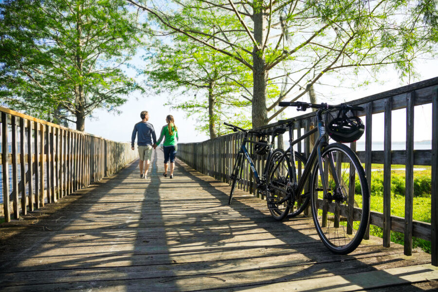 Hidden History in the Southβs Most Beautiful State Parks - 12 Two people walking on a wooden bridge toward the water. They are holding hands. Bicycles rest nearby.