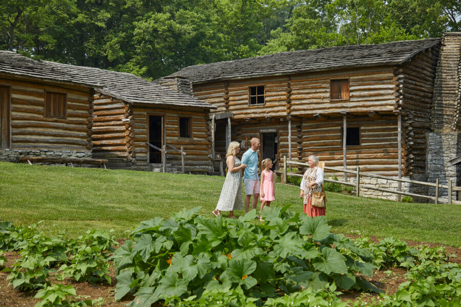 Hidden History in the Southβs Most Beautiful State Parks - 6 Four people stand and talk on grass in front of historic log cabins at a Southern state park.