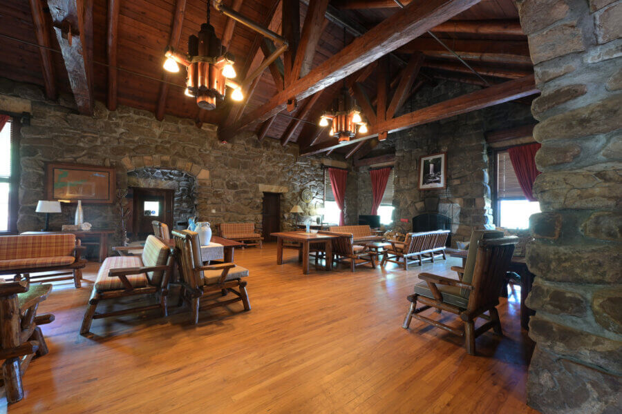 Hidden History in the Southβs Most Beautiful State Parks - 4 A rustic lodge room with stone walls, exposed wooden beams, hardwood floors, and wooden furniture arranged in seating areas under large windows with red curtains.