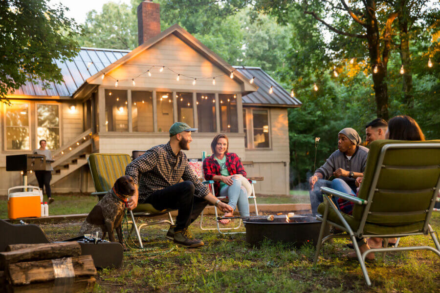 Hidden History in the Southβs Most Beautiful State Parks - 7 Five people sit around a fire pit in a backyard, talking and smiling. String lights and a house are visible in the background.