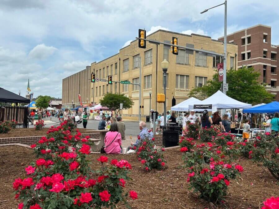 16 Can't-Miss Events in Meridian, MS This Season - 3 People visit outdoor market stalls along a street lined with blooming rose bushes, with a large beige building and traffic signals in the backgroundβa lively scene to experience on your Meridian MS things to do list.