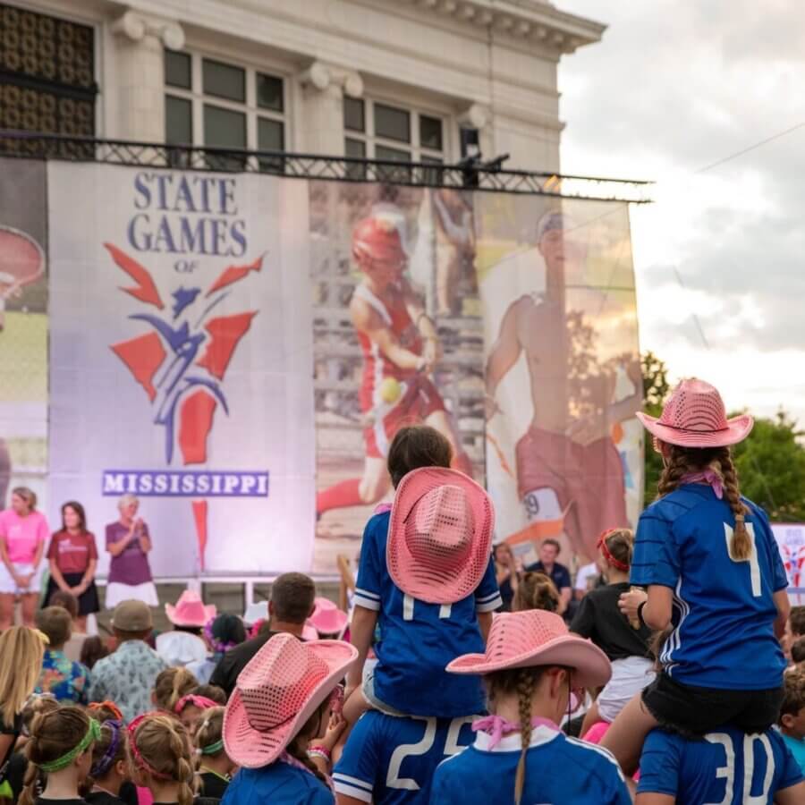 16 Can't-Miss Events in Meridian, MS This Season - 7 Children in blue jerseys and pink hats sit on shoulders in a crowd at the State Games of Mississippi eventβone of the top Meridian MS events this seasonβwith a stage and large banner in the background.