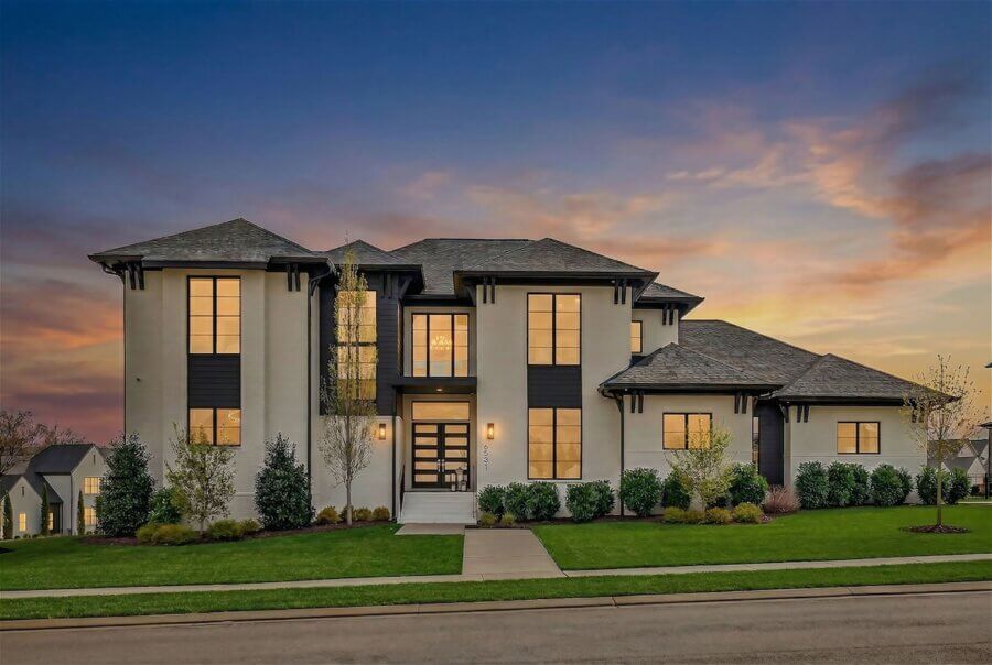 A large, modern two-story Custom Williamson County Home with beige and dark trim, illuminated windows, manicured lawn, and inviting spaces for seamless indoor living, photographed beautifully at sunset.