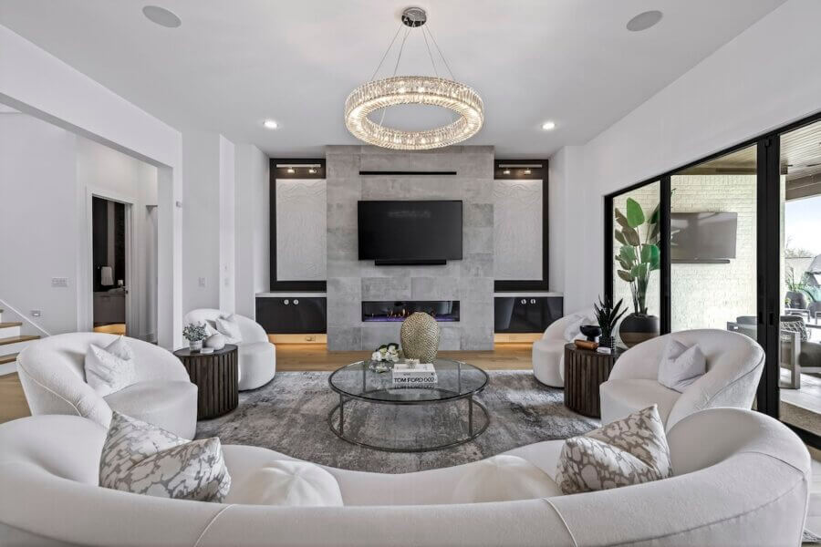 Just Listed! A Custom Williamson County Home Where Indoor & Outdoor Living Shine - 8 Modern living room with curved white sofas, a round coffee table, and a wall-mounted TV above the fireplace, seamlessly blending indoor outdoor living in this stunning Williamson County home. A large circular chandelier completes the look.