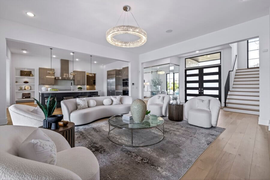 Just Listed! A Custom Williamson County Home Where Indoor & Outdoor Living Shine - 5 Modern living room in a custom home with curved white sofas, glass coffee table, neutral decor, open kitchen, and wooden stairs by a black front door. Large chandelier hangs from the ceiling for seamless indoor outdoor living.