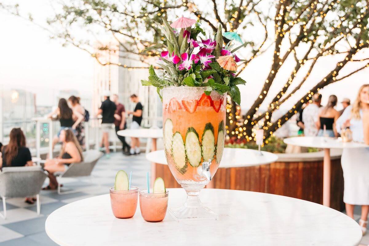 A large, decorative cocktail garnished with flowers, umbrellas, and mint sits on a marble table next to two smaller drinks in the Gulch; people socialize in the background on a rooftop terrace at sunset.