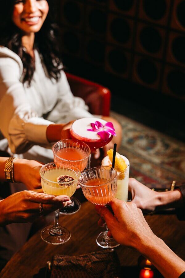 Nashville Steals, Deals & Announcements: April 2026 - 5 Four people clink cocktail glasses in a celebratory toast at a dimly lit bar; one drink is garnished with a purple flower, setting the scene for unforgettable nights and exclusive Nashville deals this April 2026.