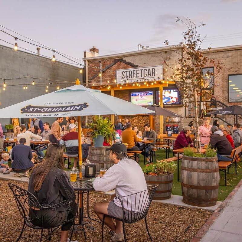 Nashville Steals, Deals & Announcements: April 2026 - 2 Outdoor patio at Streetcar bar, where people enjoy eating and drinking under umbrellas and string lights, while catching the game on a large TV—discover the latest Nashville deals in this lively setting.