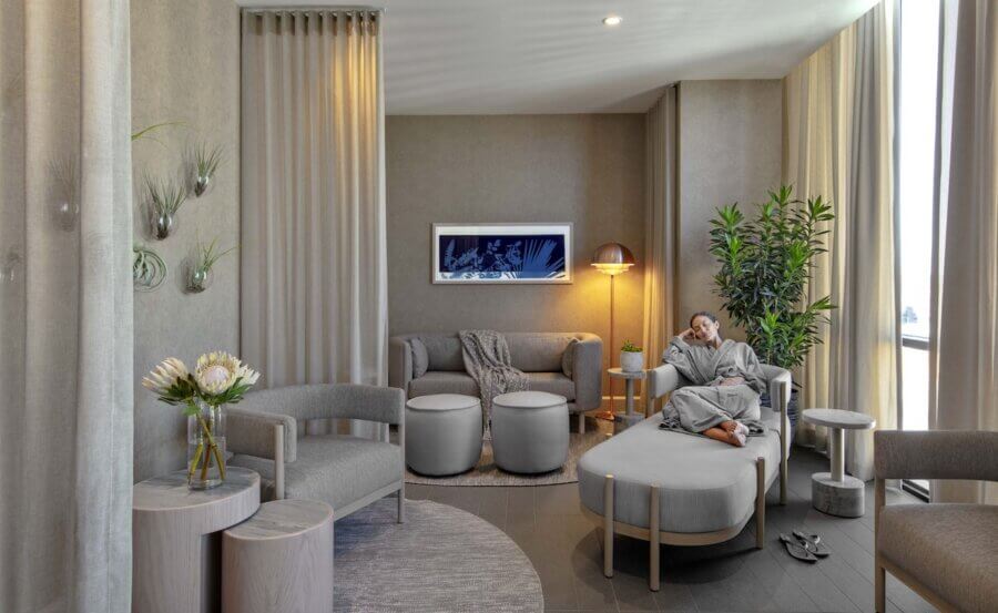 Nashville Steals, Deals & Announcements: April 2026 - 6 A woman in a robe sits on an ottoman in a modern, neutral-toned lounge with contemporary furniture, potted plants, and wall art—perfect for those seeking Nashville Deals on stylish interiors.