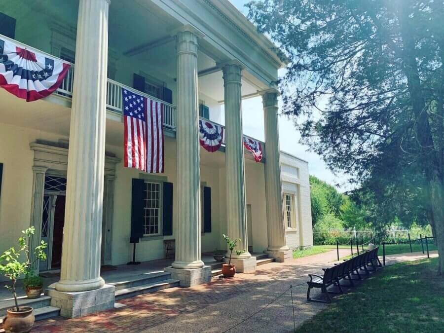 Nashville Steals, Deals & Announcements: April 2026 - 9 A historic building with tall columns is decorated with U.S. flags and patriotic bunting. Benches line the walkway, and trees provide shade on a sunny day—perfect for catching up on the latest Nashville Announcements.