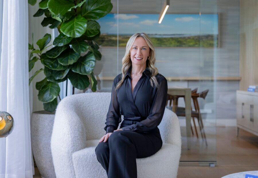 Nashville Steals, Deals & Announcements: April 2026 - 8 A woman in a black outfit sits on a white armchair in a modern office with a large plant and a glass wall behind her, preparing Nashville announcements for April 2026.