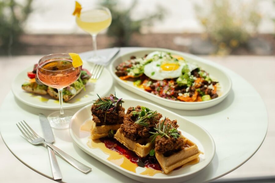 Your Updated Guide to The Gulch, One of Nashville's Most Bustling Neighborhoods - 5 Three plates of food and two cocktails on a table in the Gulch; dishes include chicken and waffles, a salad with a fried egg, and toast topped with vegetables.