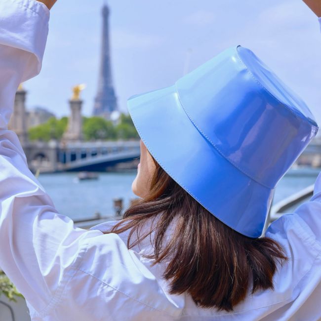 A person in a shiny blue bucket hat and white shirt, ready for April showers in chic rainwear, faces away from the camera with arms raised near the Seine River and the Eiffel Tower in the background.