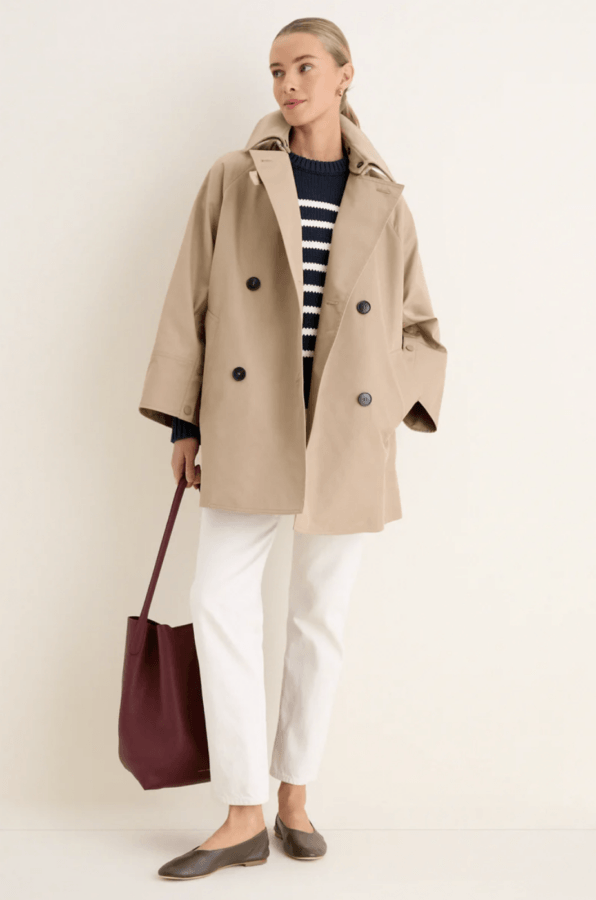 Styled for April showers, a woman stands against a light background wearing a beige double-breasted coat, striped sweater, white pants, brown shoes, and carrying a burgundy tote bag.