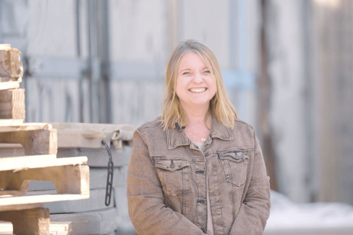 Karen Bell, with blonde hair and a warm smile, stands outside in a brown denim jacket. Wooden pallets and industrial materials set the scene behind her in this candid FACES portrait.