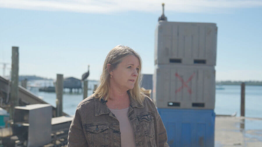 Meet the Woman Keeping Florida’s Oldest Fishing Village Thriving - 4 Karen Bell, a woman with blonde hair wearing a brown jacket, stands outdoors near stacked crates by the water, with a bird perched in the background.