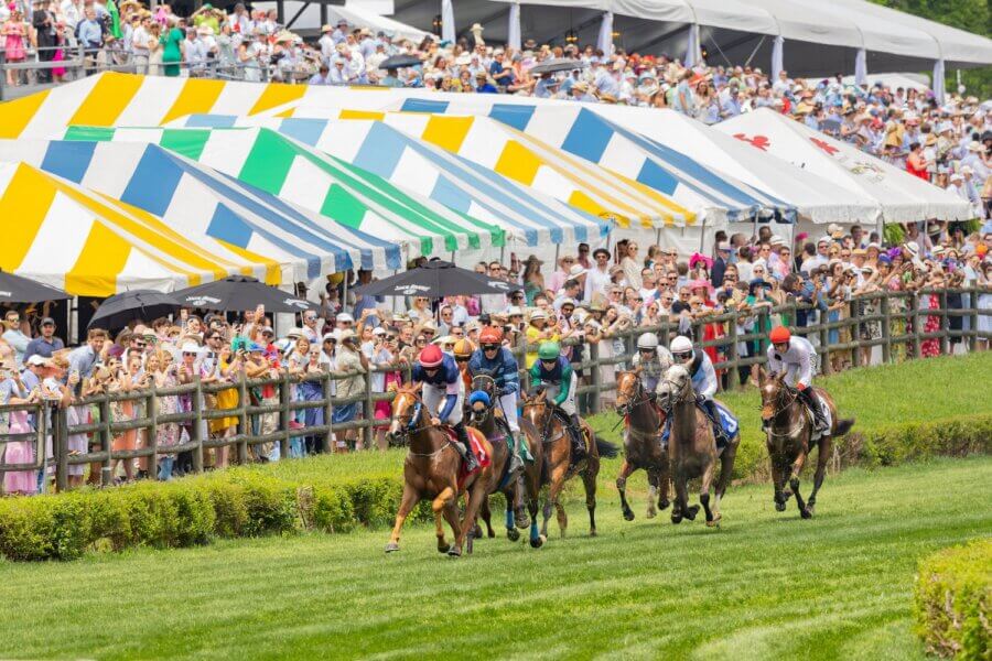Thoroughbred Horse Races Worth Traveling For - 3 Jockeys riding horses race on a grassy track at one of the top horse races, with colorful striped tents and a large crowd of spectators in the background.