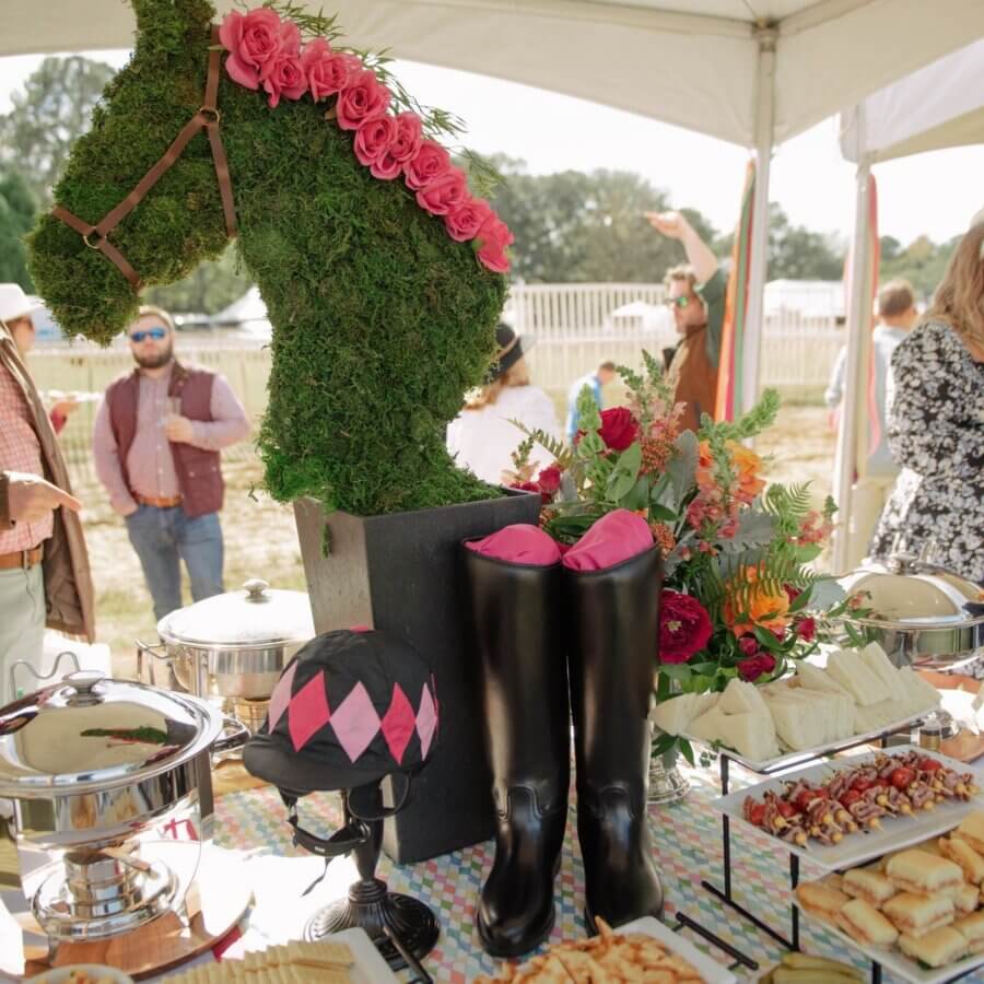 Thoroughbred Horse Races Worth Traveling For - 4 Buffet table with an equestrian-themed centerpiece, featuring a moss horse head with pink roses, black riding boots with pink inserts, a riding helmet, and assorted food trays—perfect for celebrating the excitement of thoroughbred horse races.
